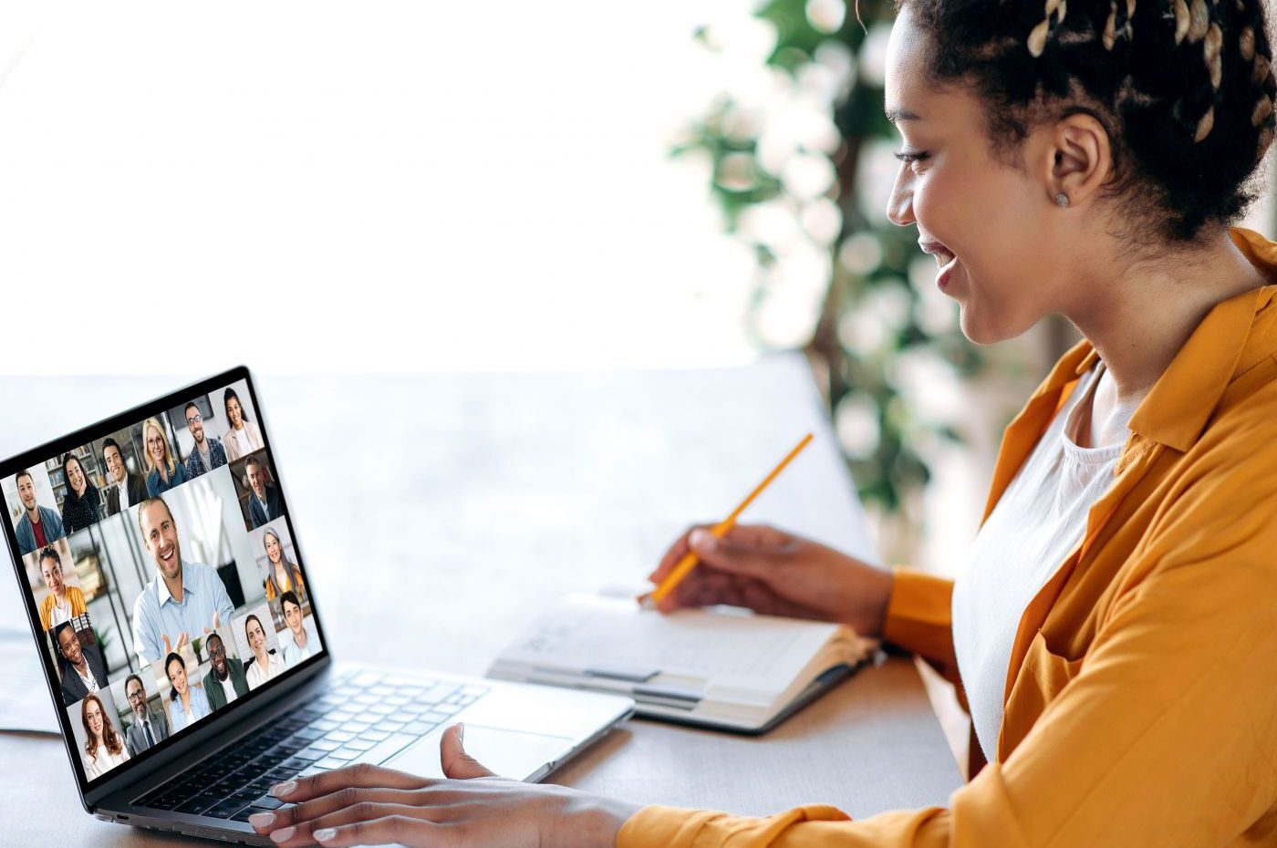 Frau sitzt am Laptop in der Online-Konferenz, lächelt und macht Notizen in ein Buch.