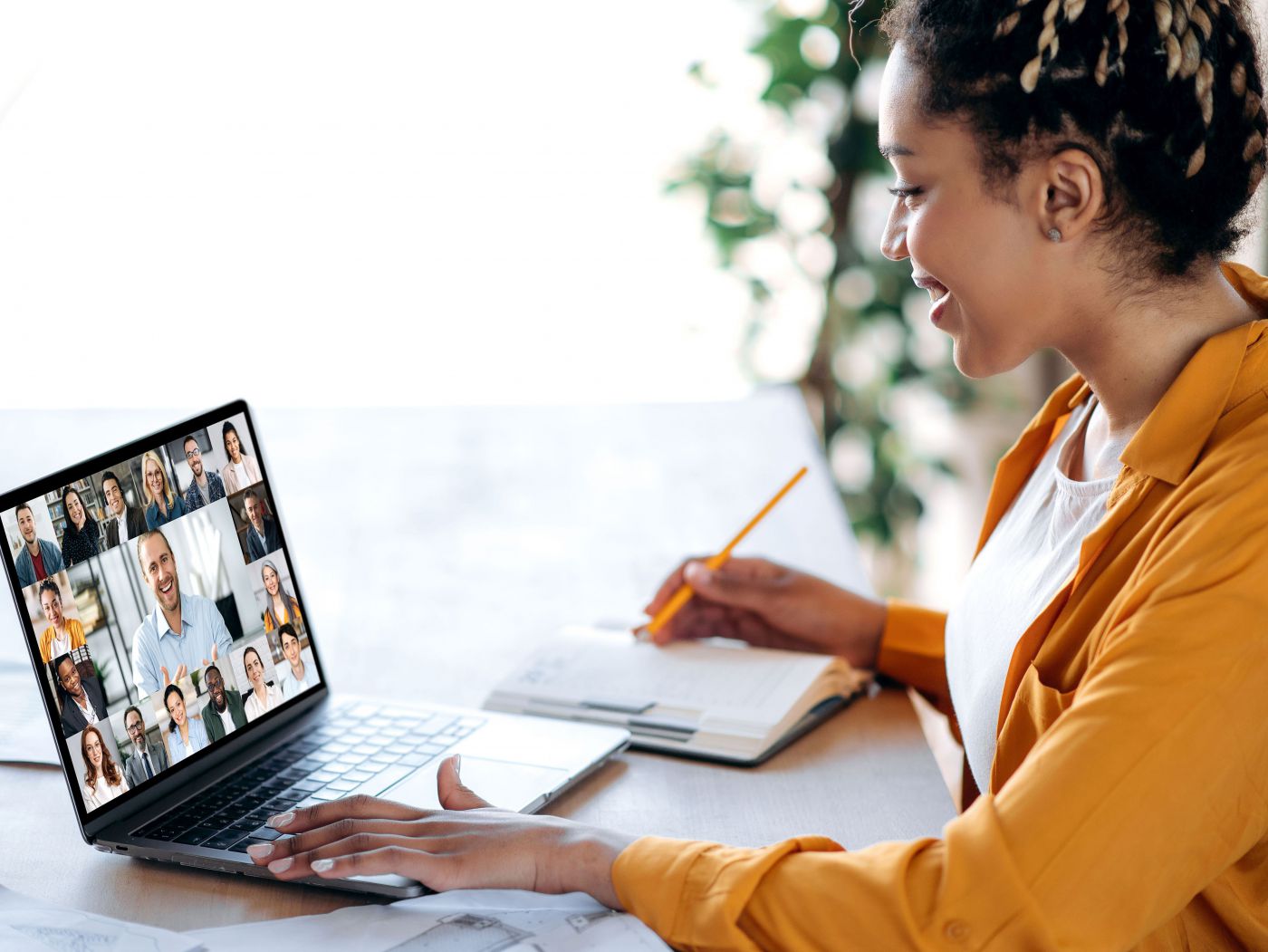 Frau sitzt am Laptop in der Online-Konferenz, lächelt und macht Notizen in ein Buch.